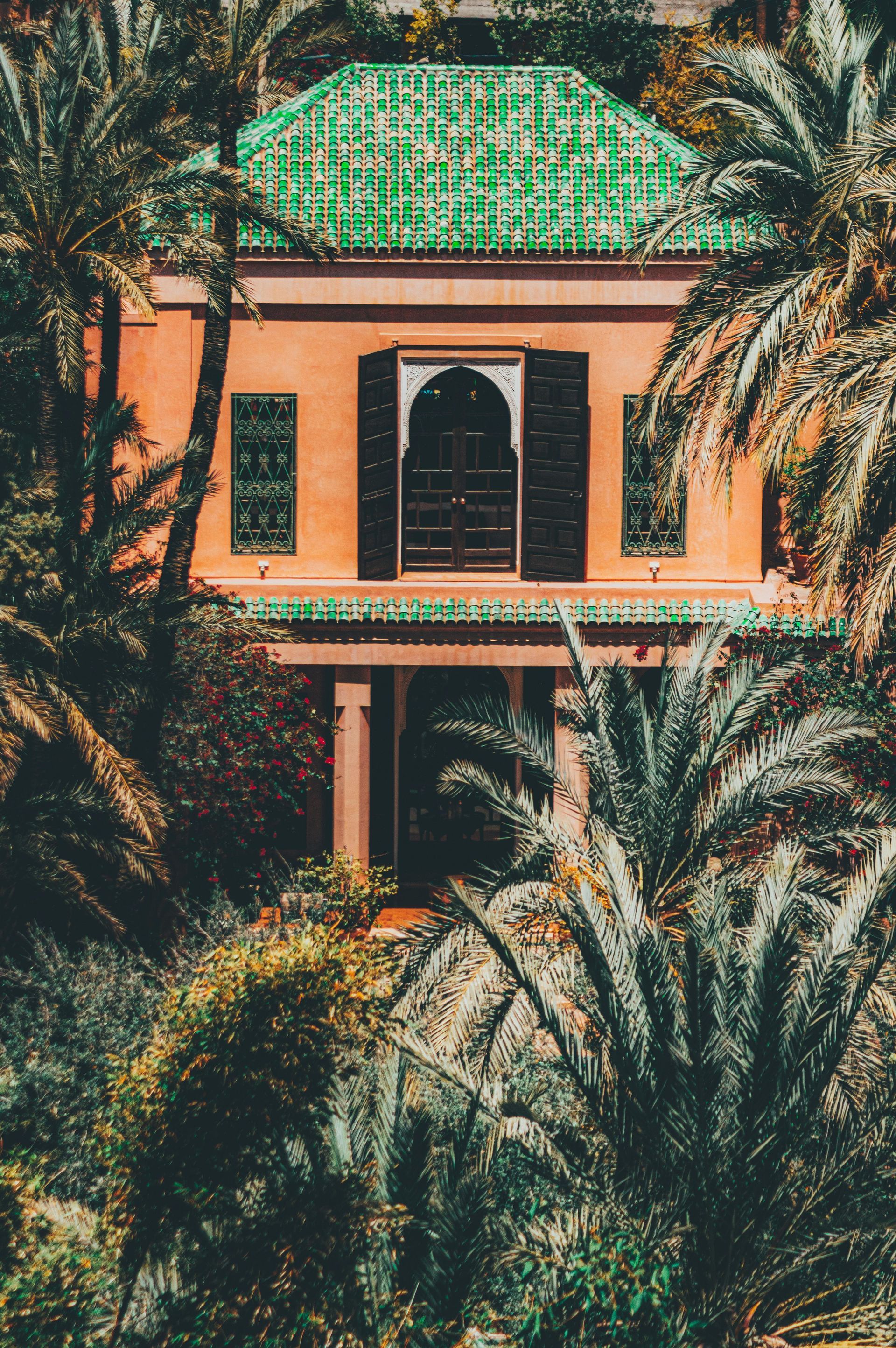 A terracotta-colored building with a green tiled roof surrounded by palm trees and lush foliage.