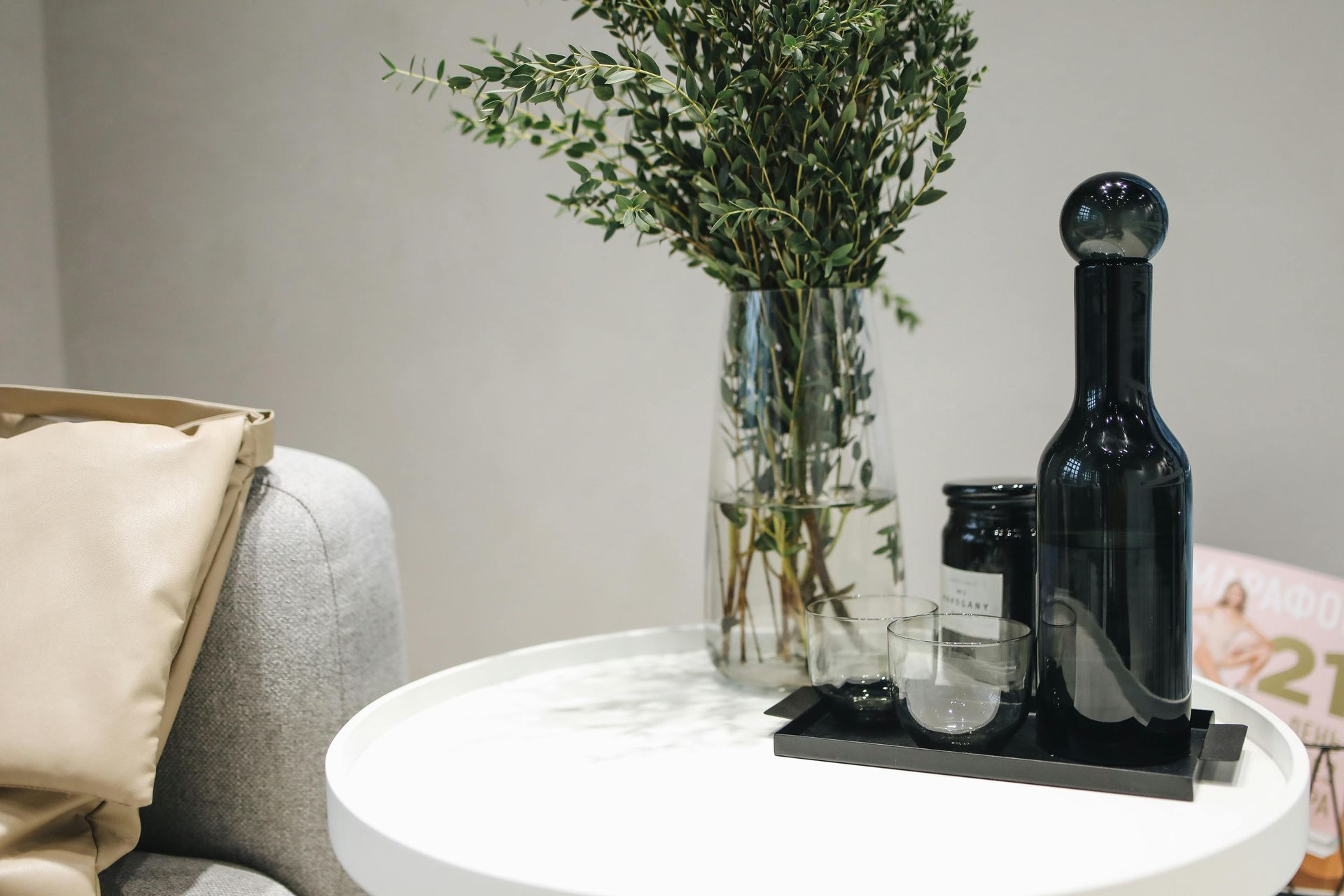 Round white table with a black bottle, tray, glass, and greenery beside a beige sofa