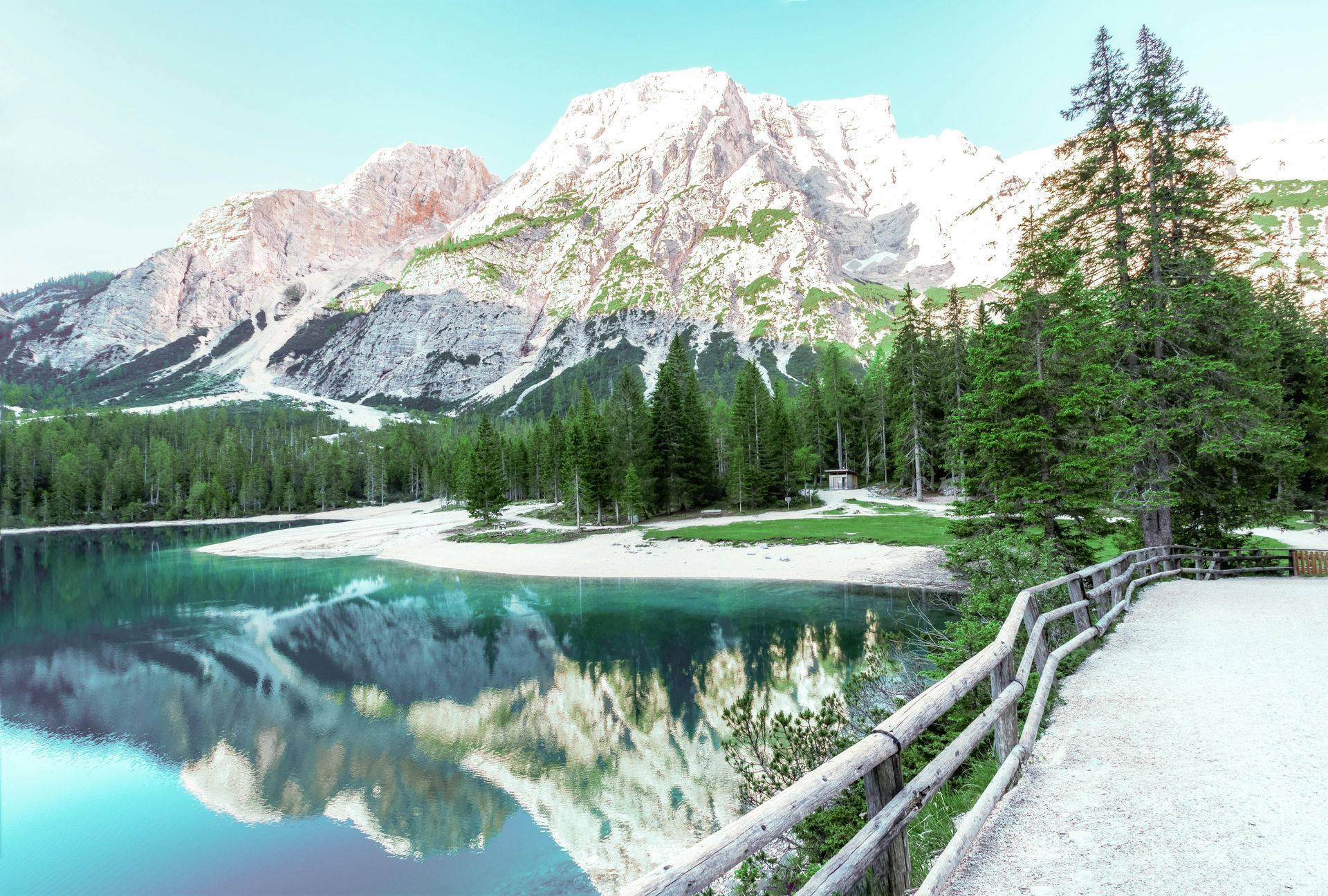 Lakeside view with mountain backdrop. Turquoise water reflects snow-capped peaks, surrounded by green trees and a wooden fence.
