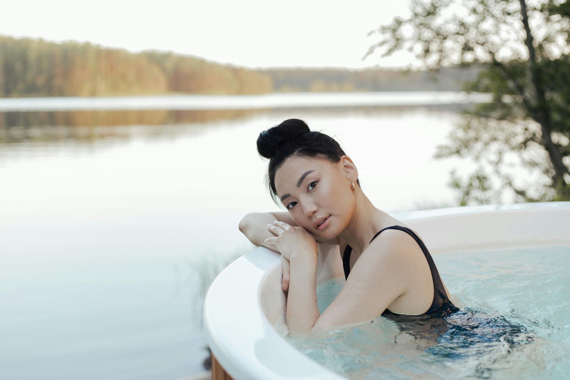 Person relaxing in a white hot tub by a calm lakeside, leaning on the rim with a serene expression