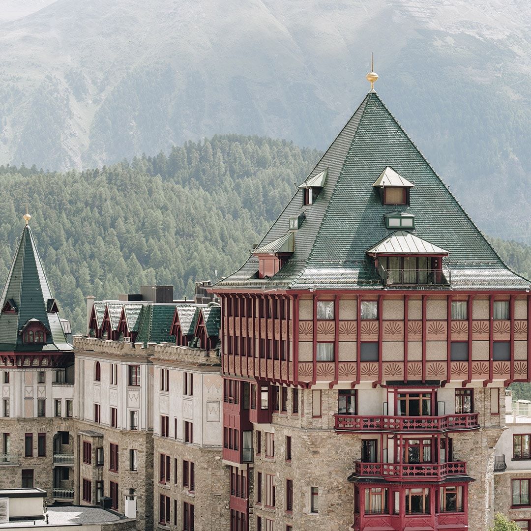 Stone building with green tiered roof, red trim, and small balconies, in a mountain setting. The Legacy of Badrutt’s Palace Hotel 