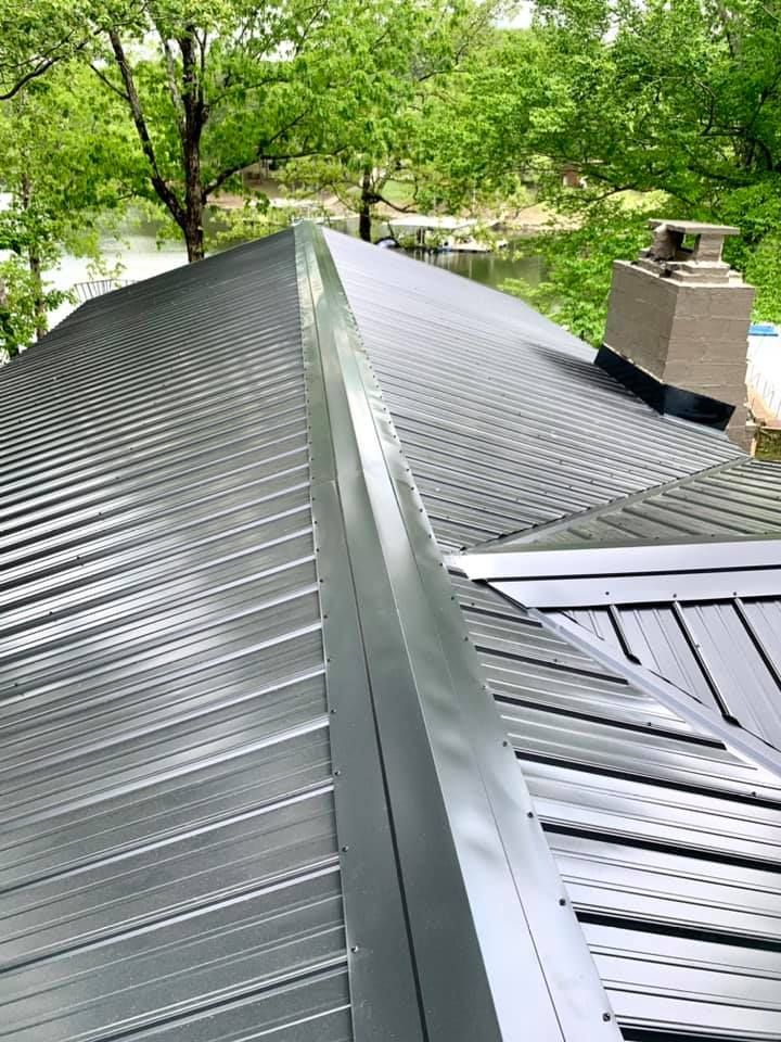 High-angle view of a dark metal roof with a central ridge cap, a stone chimney, and trees in the background.