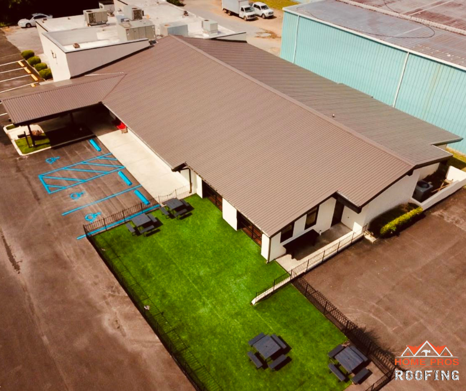 Aerial view of a commercial building with a brown metal roof, adjacent parking lot, and a small green lawn with tables.