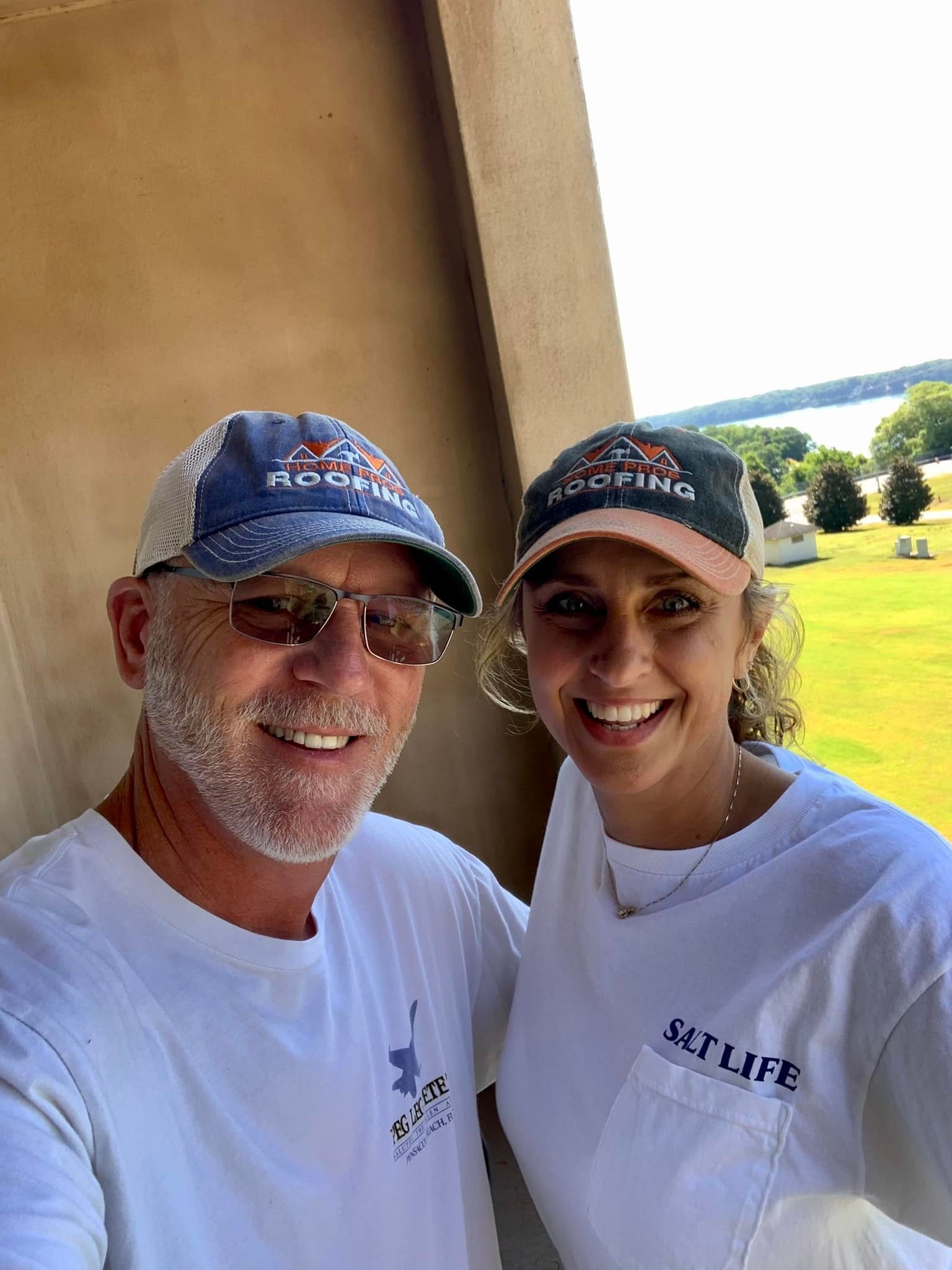 A smiling couple in matching white shirts and branded baseball caps standing outdoors in front of a sunny, grassy view.