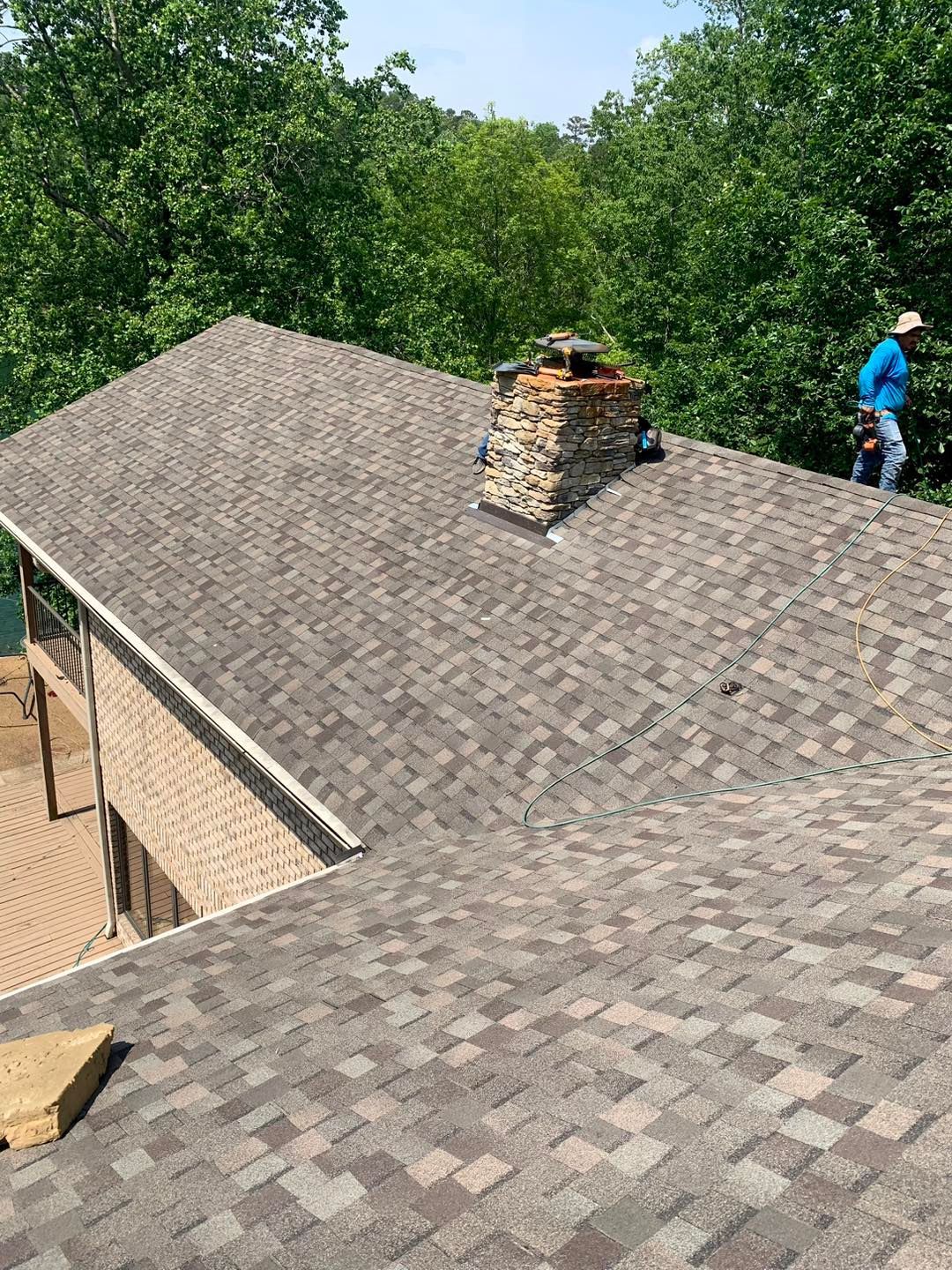 A worker in a blue shirt repairs a textured stone chimney on a residential shingled roof surrounded by green trees.