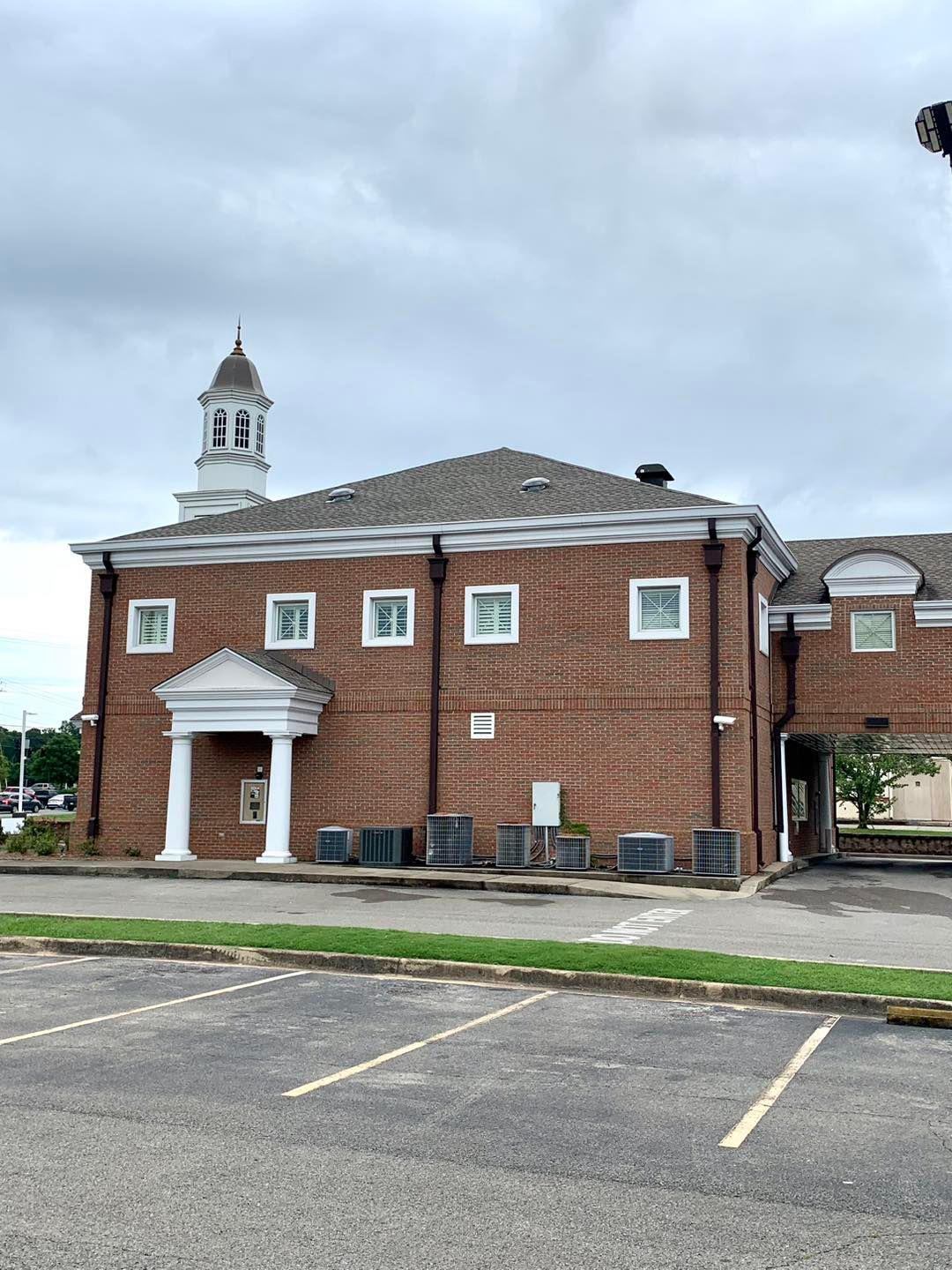 A two-story brick building with a white columned entrance, a cupola on the roof, and a parking lot in the foreground.