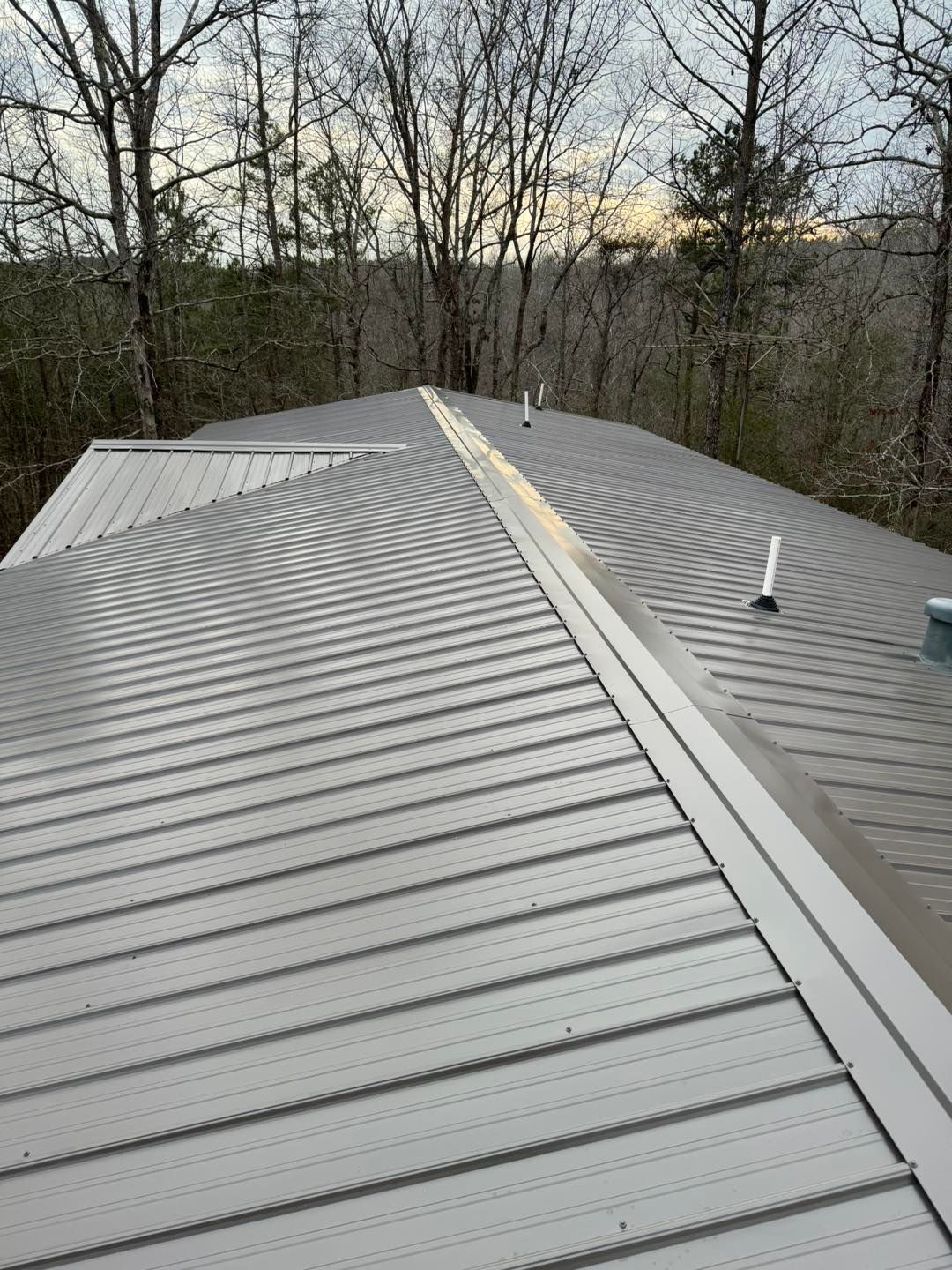 A high-angle view of a gray metal roof with a central ridge cap and two white plumbing vents, overlooking a forest.