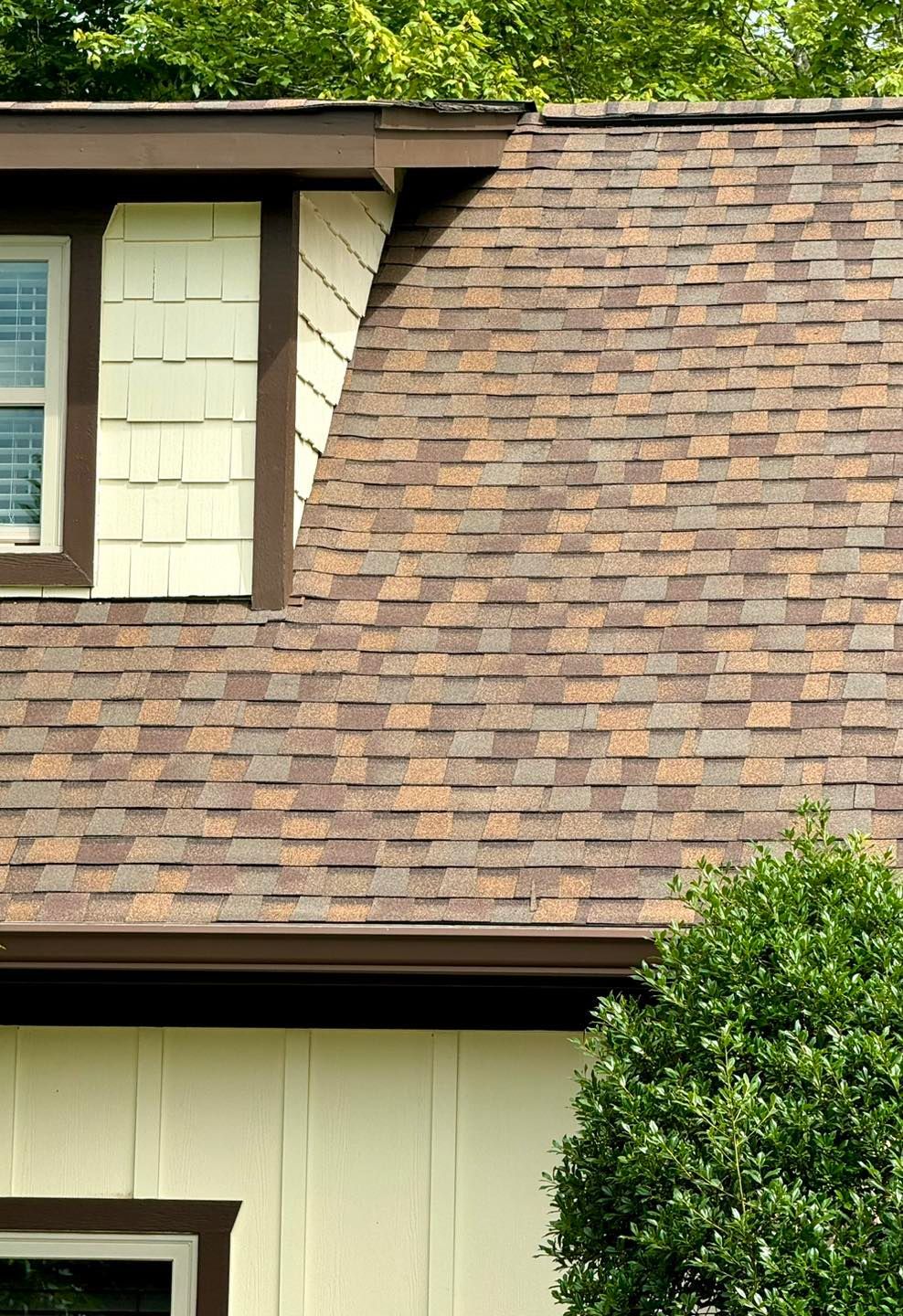 A dormer window with cream-colored shingle siding sits on a house roof covered in brown and tan architectural shingles.