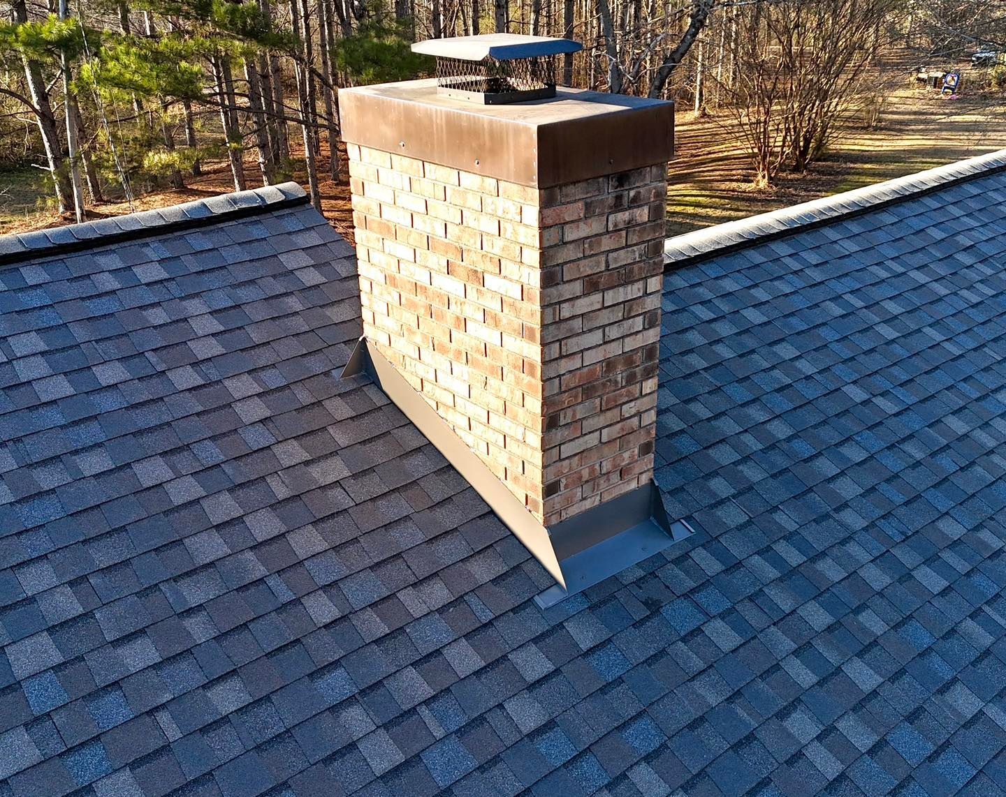 A brick chimney with a metal cap and flashing sits on a dark shingled roof, set against a backdrop of trees.