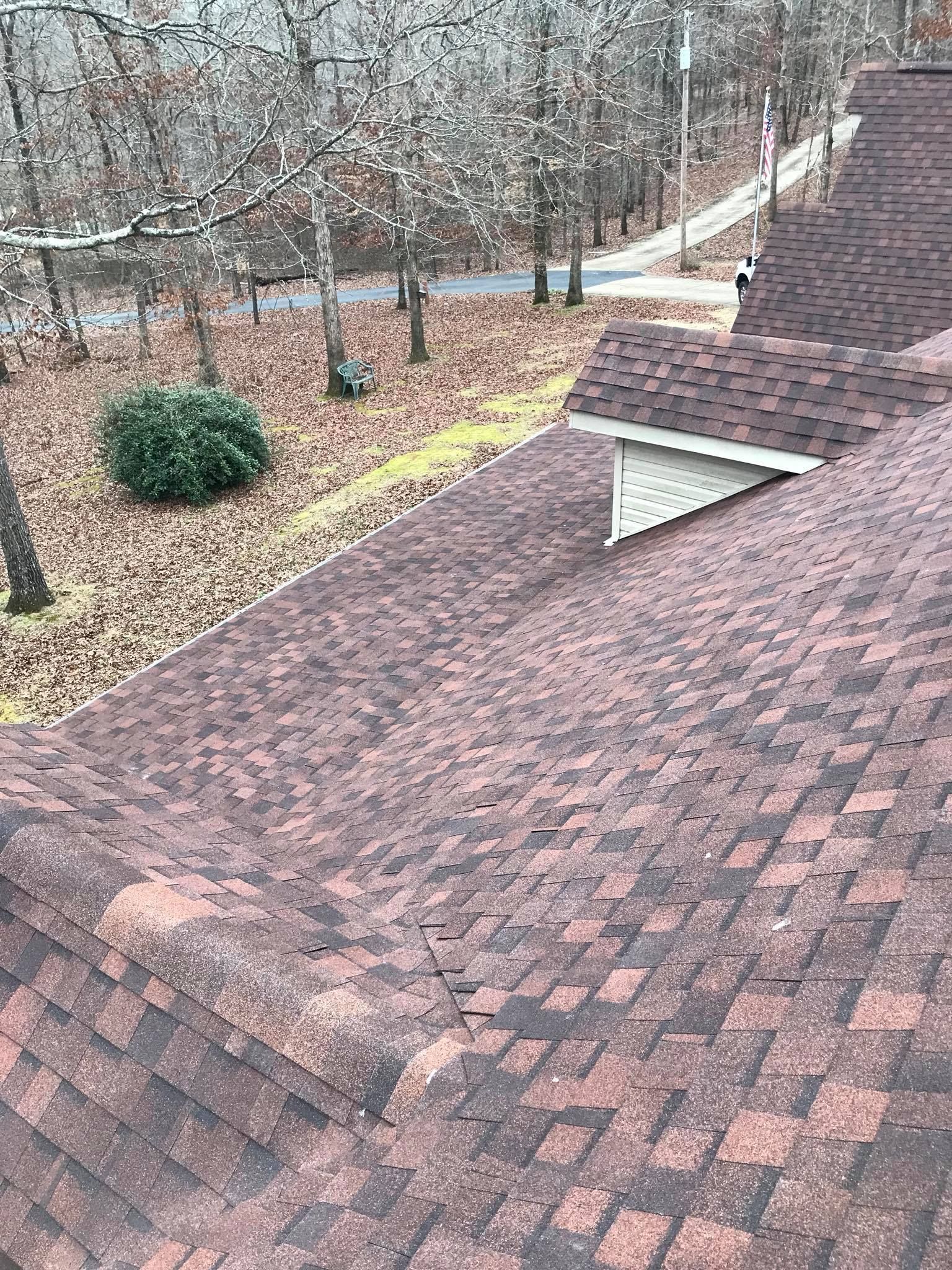 An angled view of a textured, reddish-brown shingled roof with a small dormer, overlooking a wooded area in late autumn.