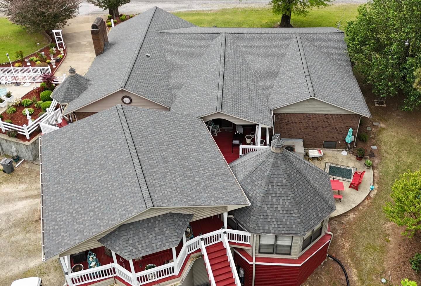 An aerial view of a brick house with a complex, multi-gabled gray shingle roof and a wrap-around porch with red railings.