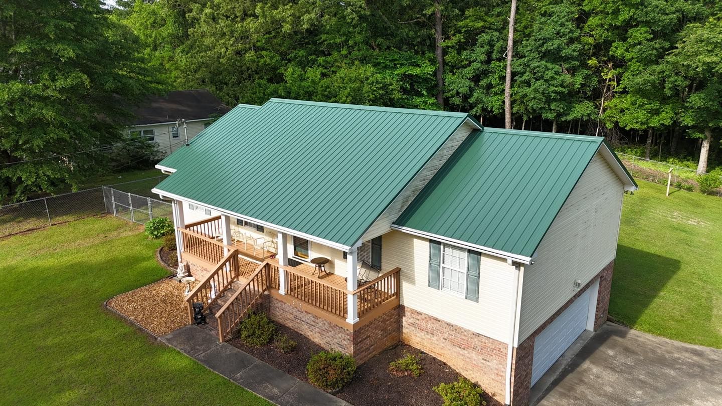 An aerial view of a suburban house with a green metal roof, a front porch, and a side-entry garage, set in a grassy yard.