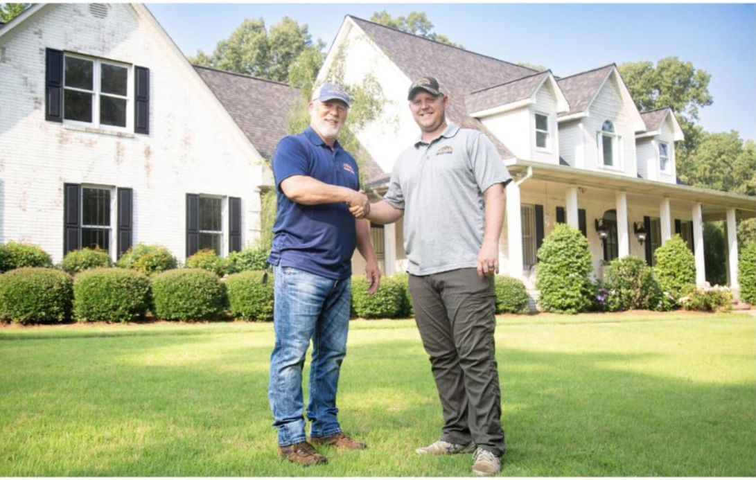 Two people shake hands on a lawn in front of a white two-story farmhouse with a covered porch and dormer windows.