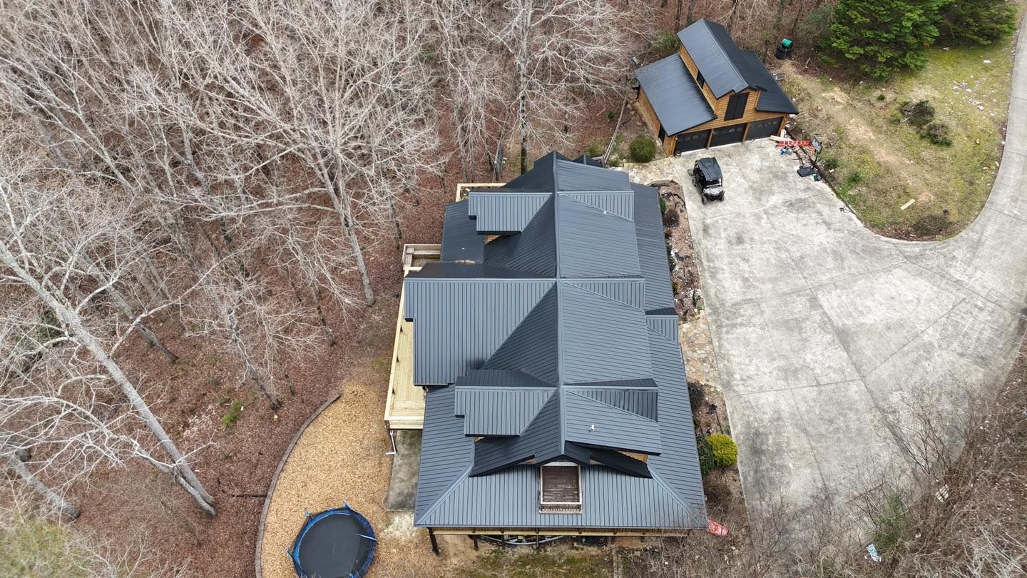 A high-angle view of a house with a dark metal roof, surrounded by bare trees, a gravel driveway, and a small yard.