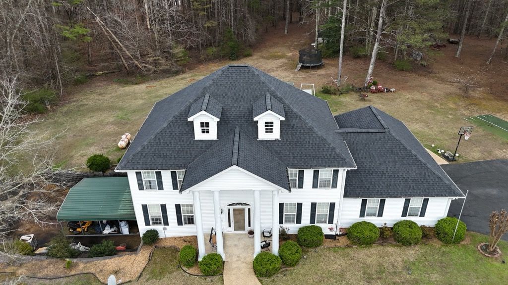 Aerial view of a large white house with black roof and dormer windows, surrounded by trees and a front lawn.