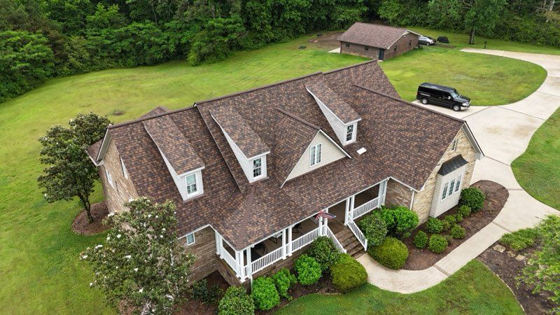 Aerial view of a large suburban house with a brown roof, white trim, and a curved driveway surrounded by green lawn.