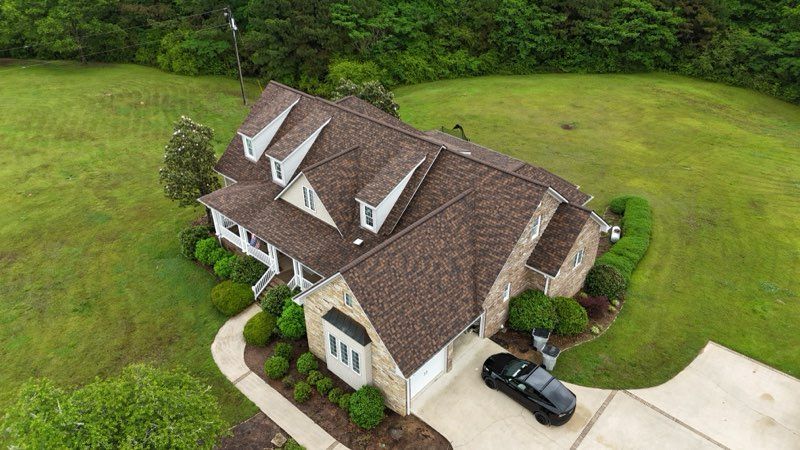 Aerial view of a large beige house with a brown roof, driveway, and green lawn surrounding it.