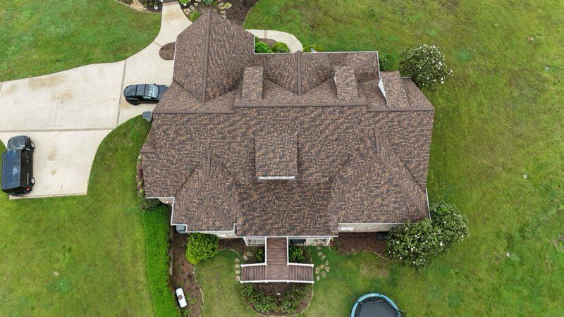 Aerial view of a brown-roofed house with driveway, cars, and green lawn.