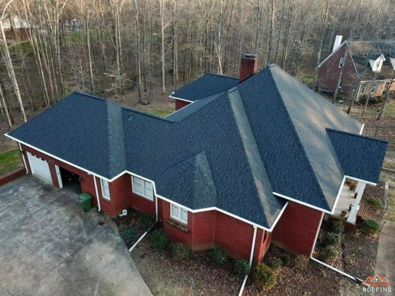 An aerial view of a single-story brick house with a dark shingled roof, surrounded by a wooded area and a paved driveway.