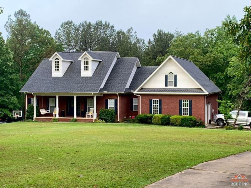 A red brick house with a covered front porch, two dormer windows, and a green lawn, surrounded by mature trees.