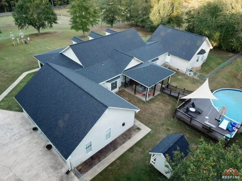 Aerial view of a white single-story home with a dark shingled roof, backyard deck, pool, and a small matching shed.