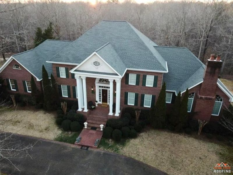 An aerial view of a two-story red brick home with a white columned porch, dark shutters, and a grey shingled roof.
