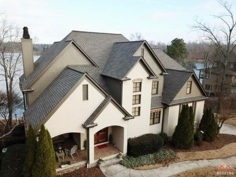 An aerial view of a tan, multi-story home with a dark shingled roof, set beside a lake and surrounded by trees.