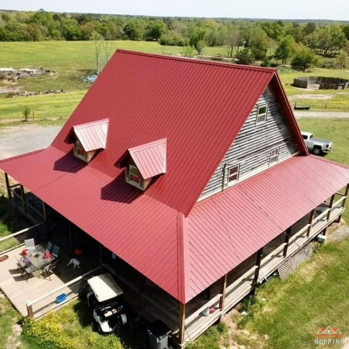 An aerial view of a rustic house with a steep, bright red metal roof, dormer windows, and a wraparound porch.