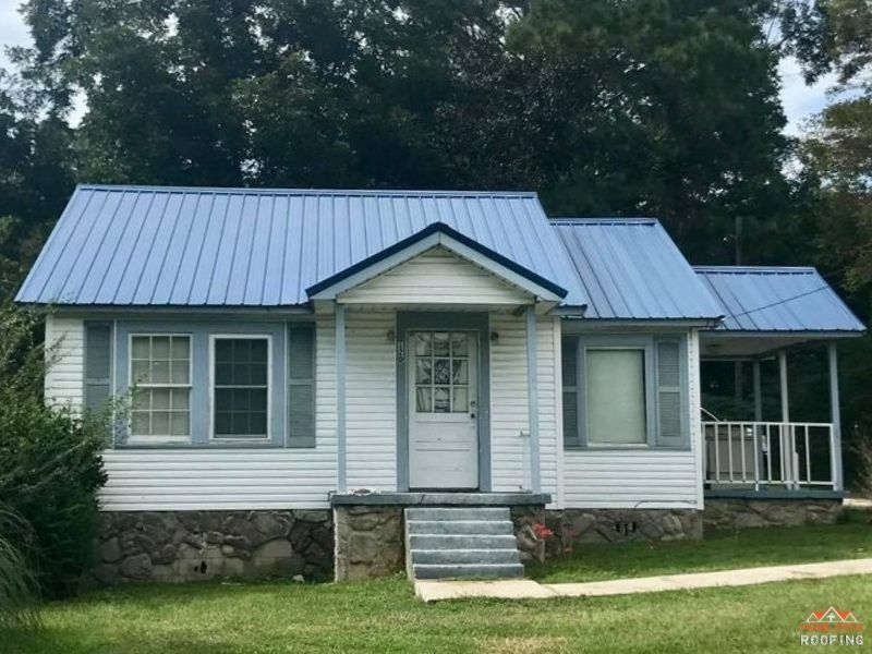 A white, single-story house with a blue metal roof, stone foundation, front porch, and a small side-covered patio.