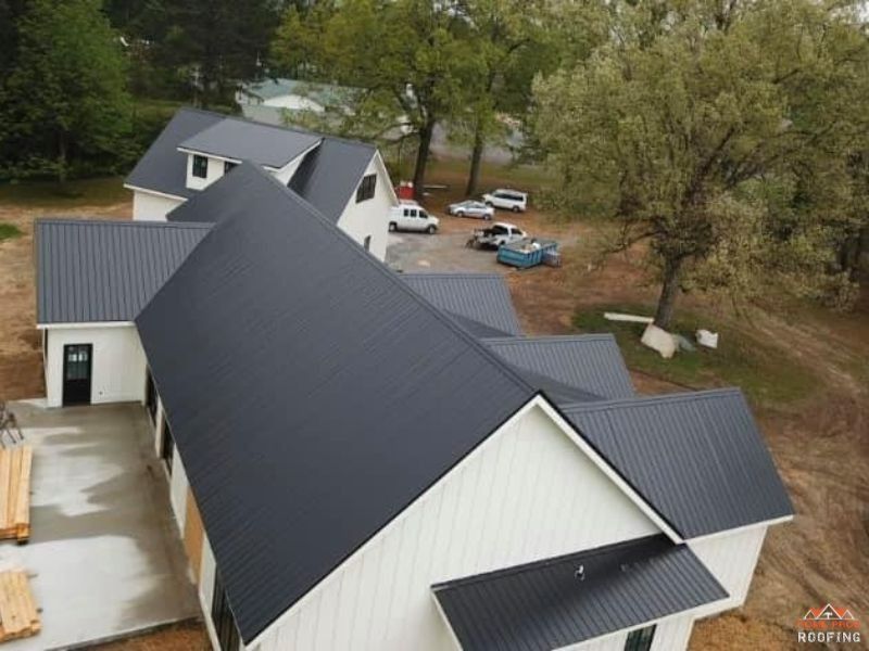 Aerial view of a white modern farmhouse with a dark metal roof, set on a wooded lot with construction vehicles nearby.