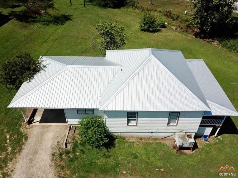 An aerial view of a white single-story house with a light-colored metal roof, surrounded by a green grassy lawn.
