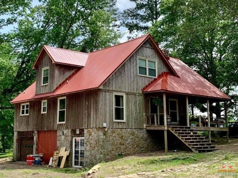 A rustic two-story house with vertical wood siding, a red metal roof, and a stone foundation, set among mature trees.