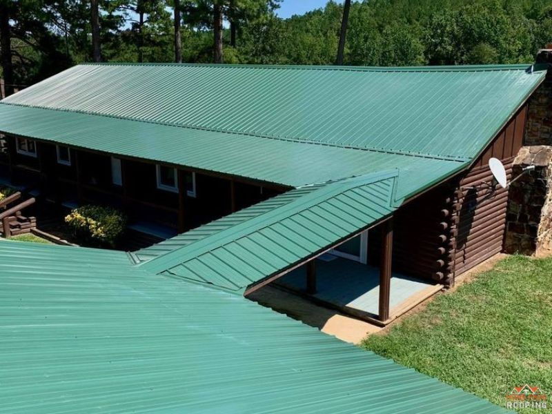 A log cabin with a prominent green metal roof and a stone chimney, surrounded by green trees and grass.