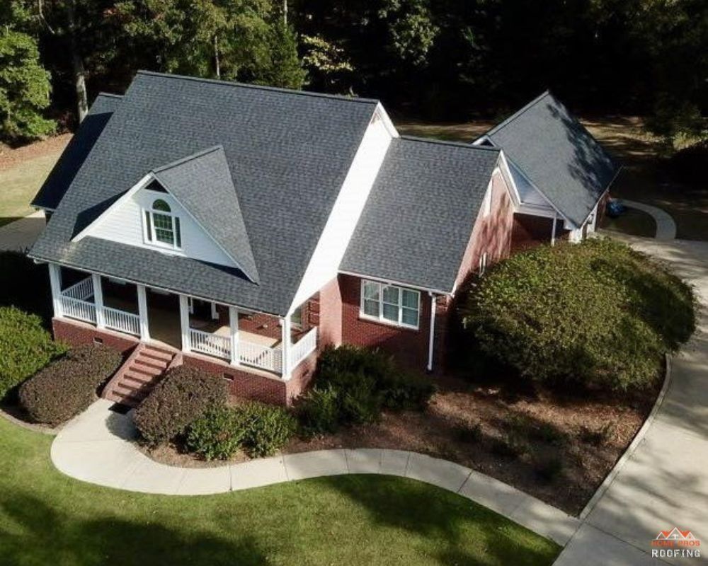 An aerial view of a brick house with a dark shingled roof, a front porch, and a curved sidewalk in a landscaped yard.