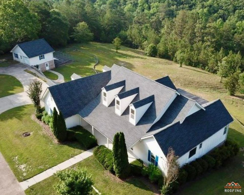 Aerial view of a white house with a dark roof, three dormer windows, and a detached garage on a grassy, wooded lot.