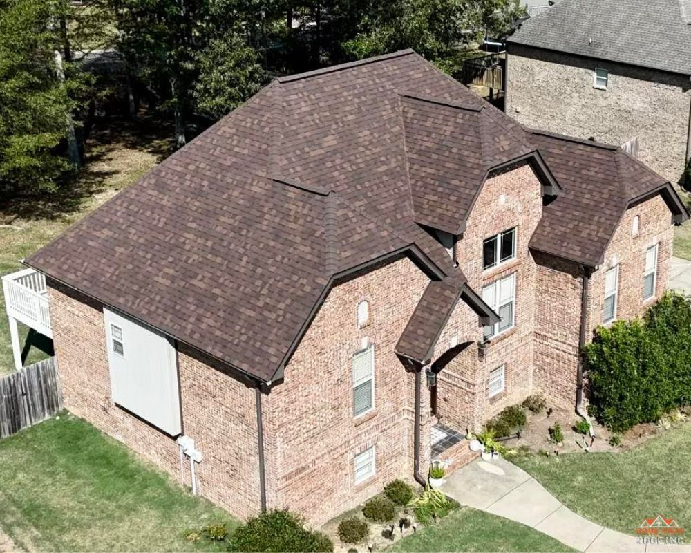 An aerial view of a two-story brick house with a dark brown shingled roof, surrounded by green grass and trees.