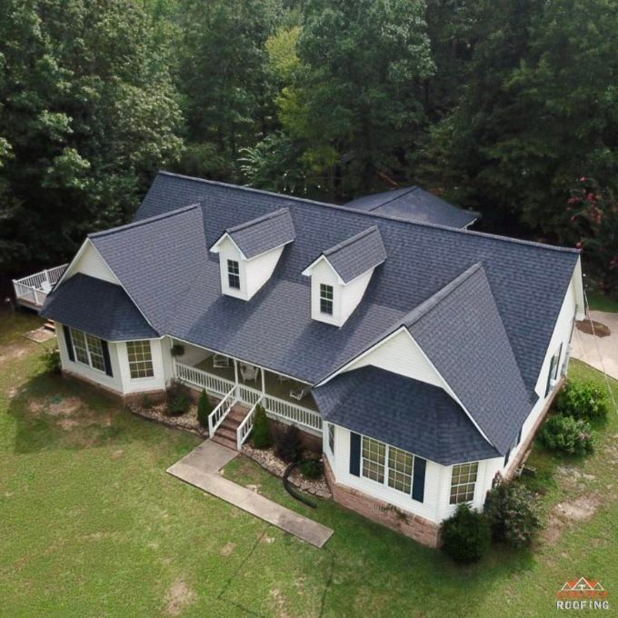 An elevated view of a white, multi-gabled house with a new, dark gray shingled roof, surrounded by a lawn and trees.