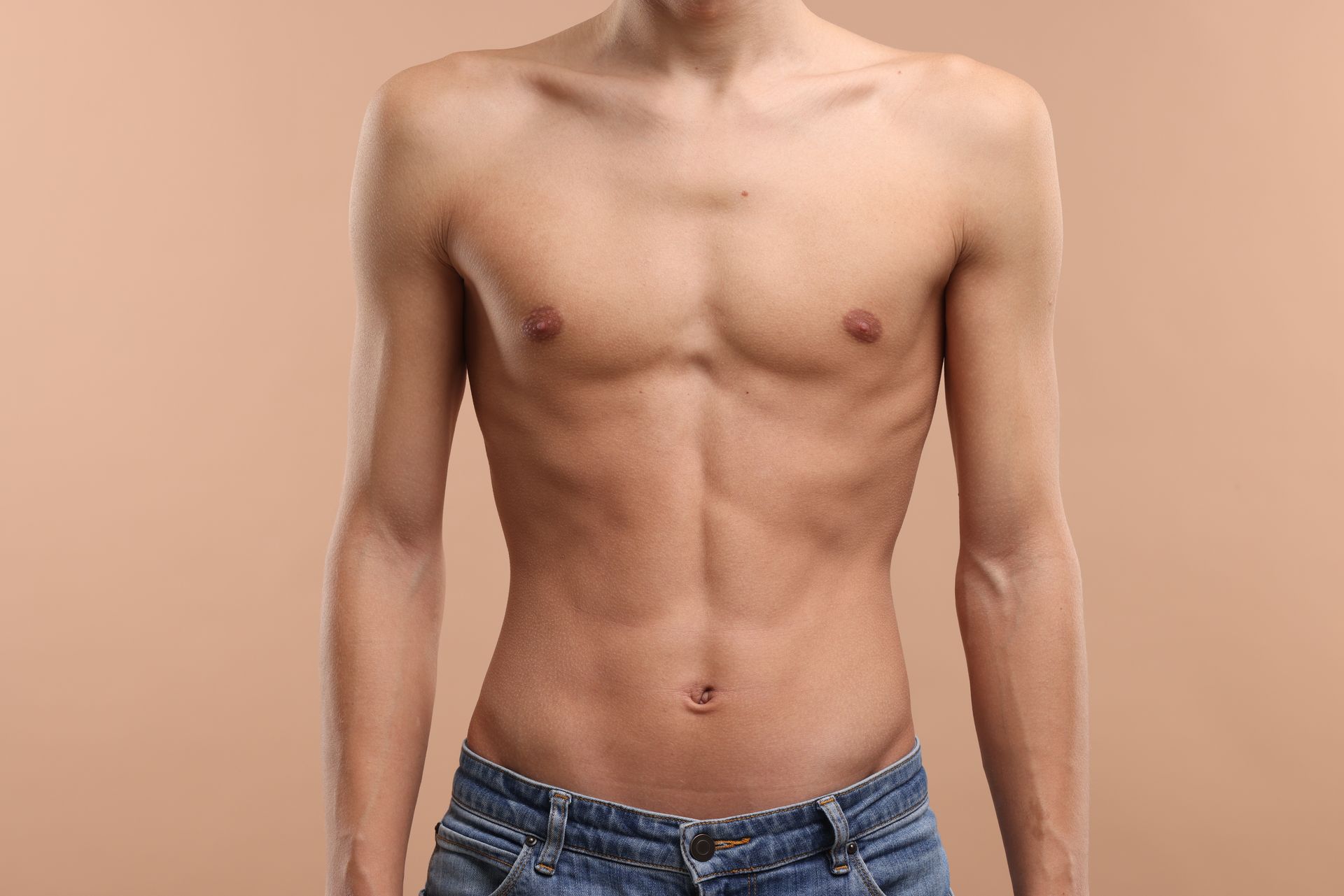 Ein Mann ohne Hemd und in Jeans steht vor einem braunen Hintergrund.