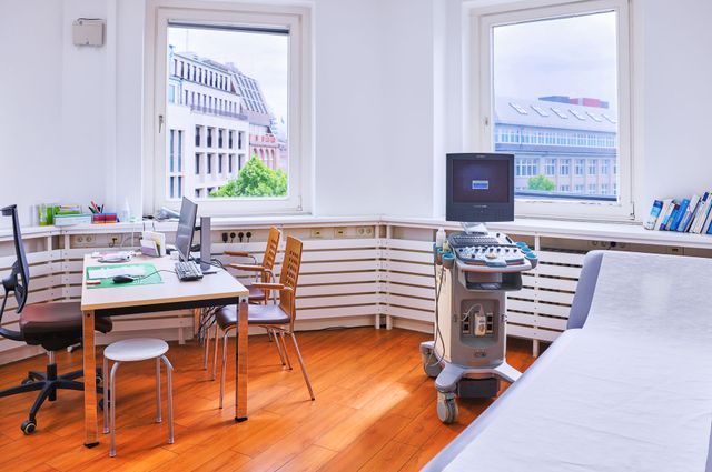 Medizinischer Untersuchungsraum mit Ultraschallgerät, Untersuchungstisch, Schreibtisch, Stühlen und Fenstern.