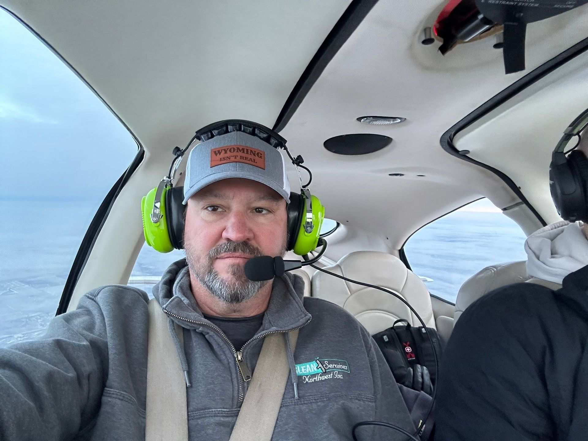 Man wearing a headset and cap seated inside a small airplane cockpit, looking at the camera.