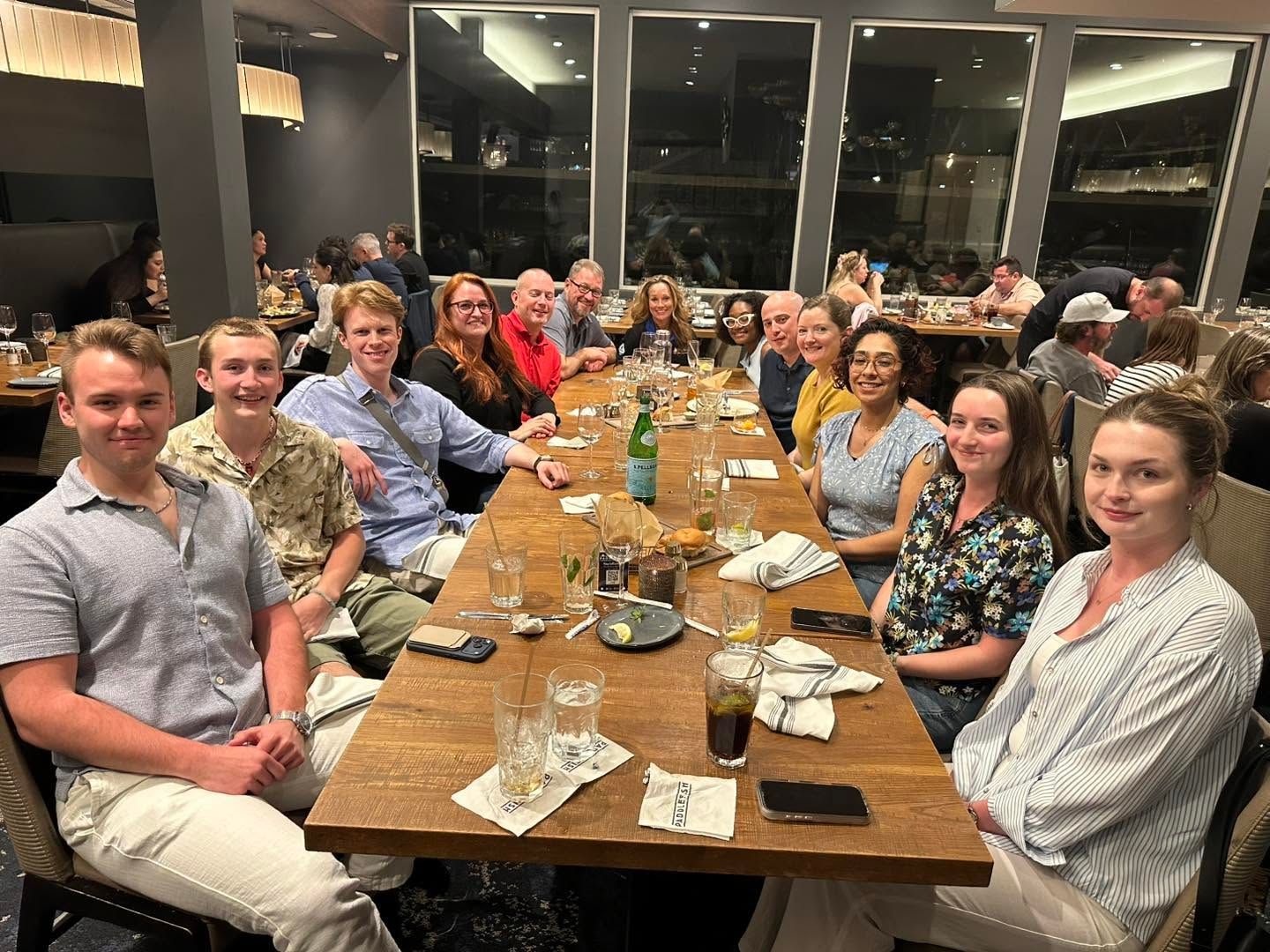 Large group seated around a long dining table at a restaurant, smiling for a meal together.