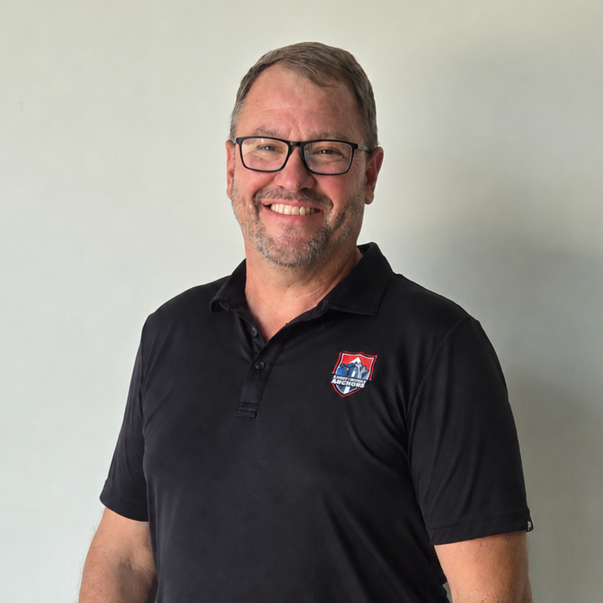 Smiling man in black polo shirt with a logo, standing against a plain light wall.