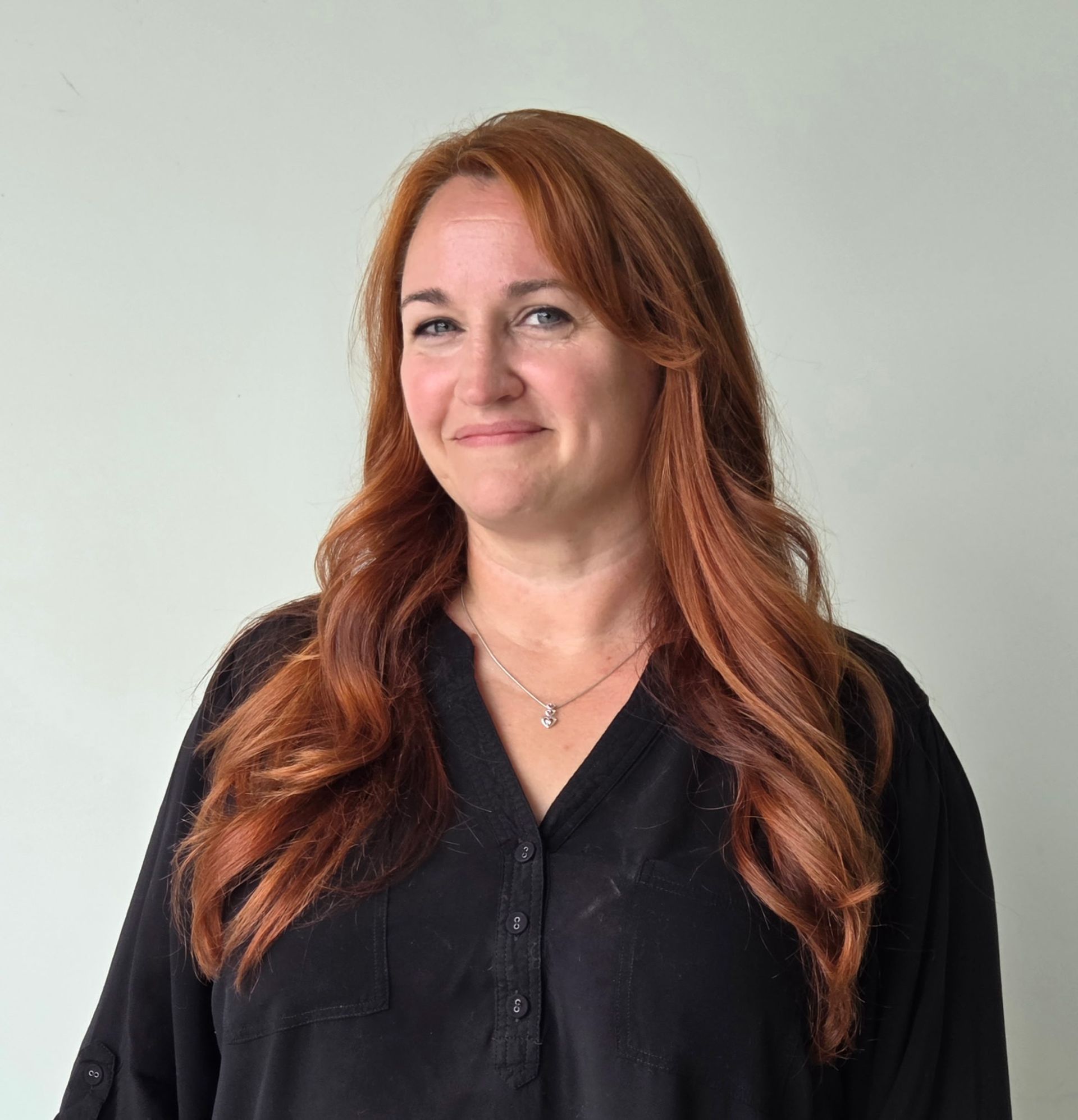 Woman with long red hair wearing a black blouse, smiling against a plain light gray background