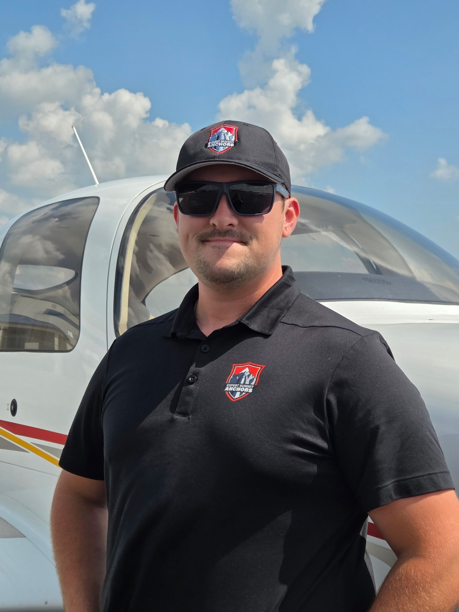 Man in black polo and cap standing by a small airplane under a blue sky