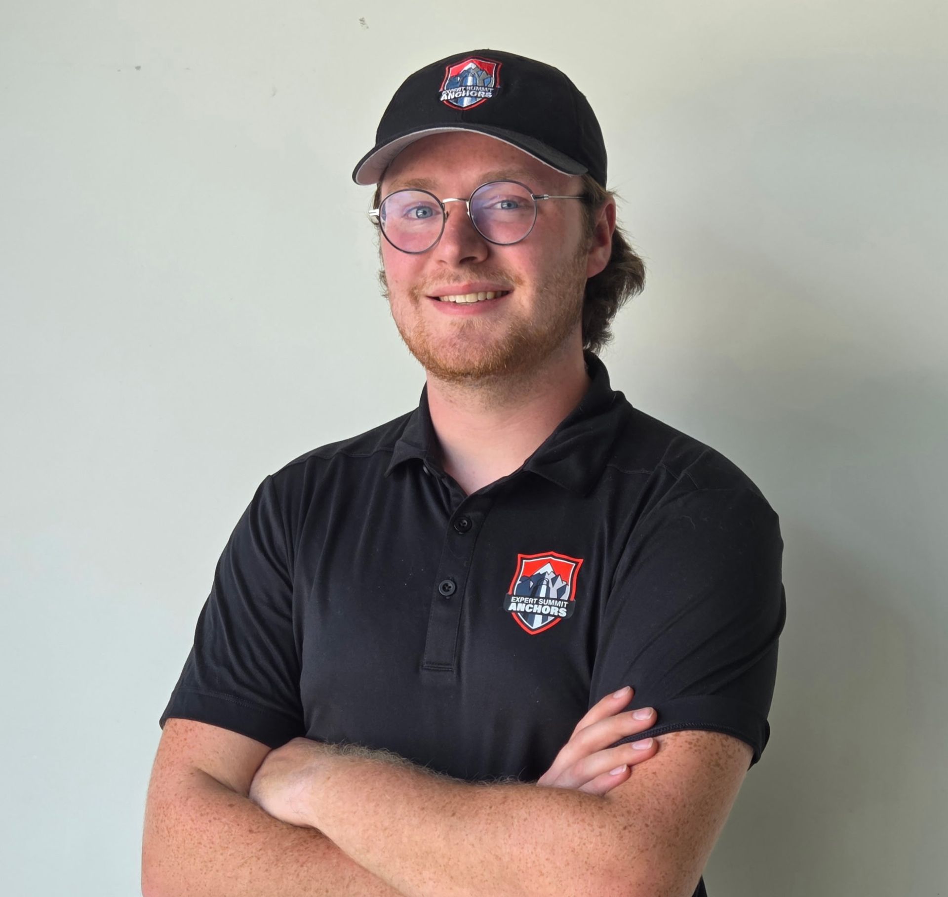 Person wearing a black cap and polo shirt, smiling with arms crossed against a plain wall.