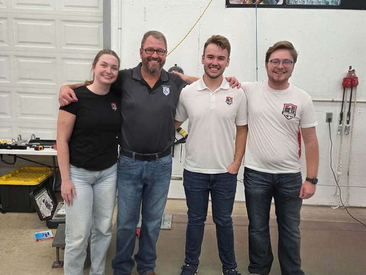 Four people smiling and standing together in a workshop or garage with tools and equipment in the background