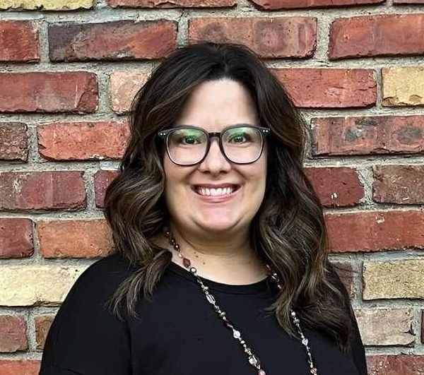 Smiling woman with glasses standing against a red brick wall