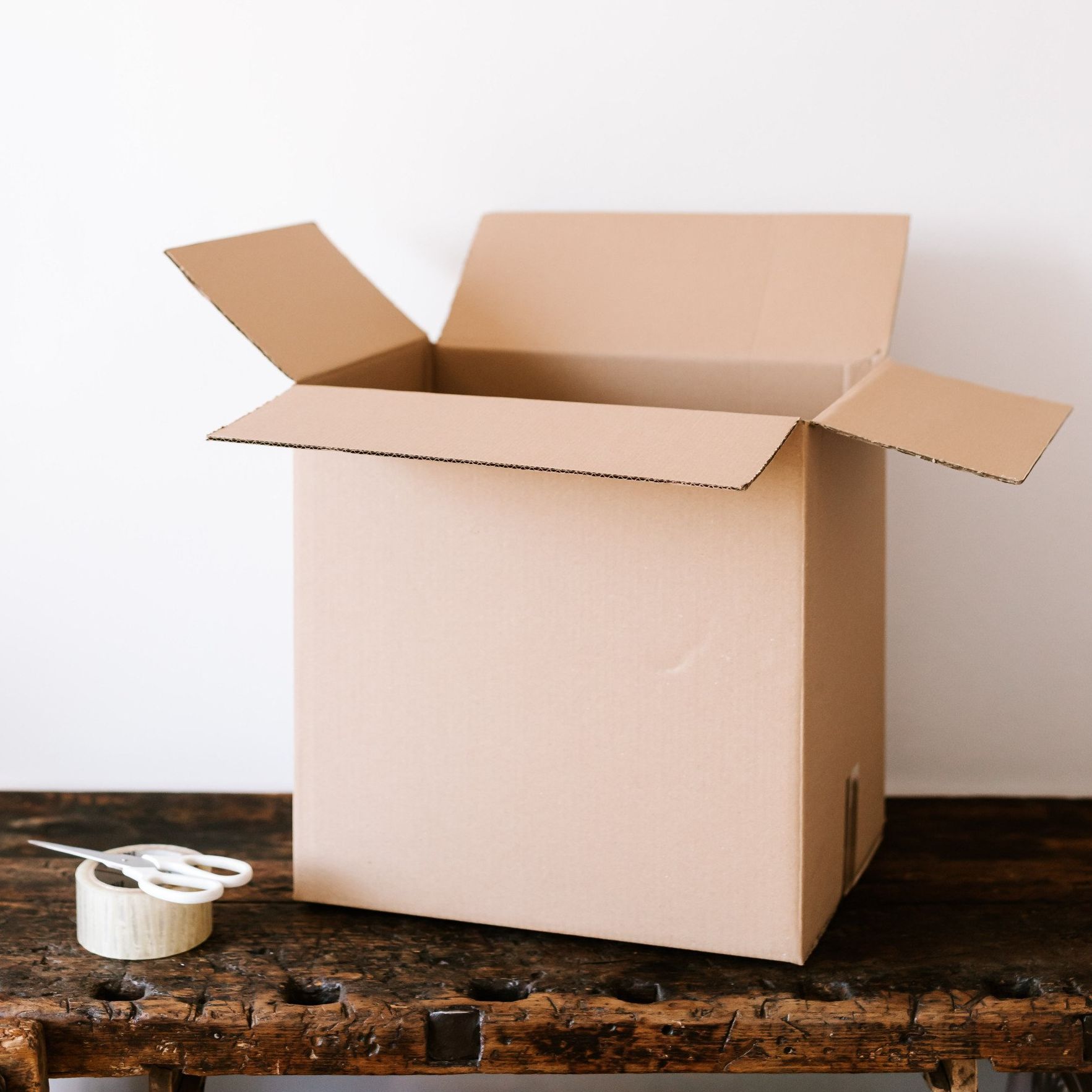 An open cardboard box is sitting on a wooden table next to a roll of tape.