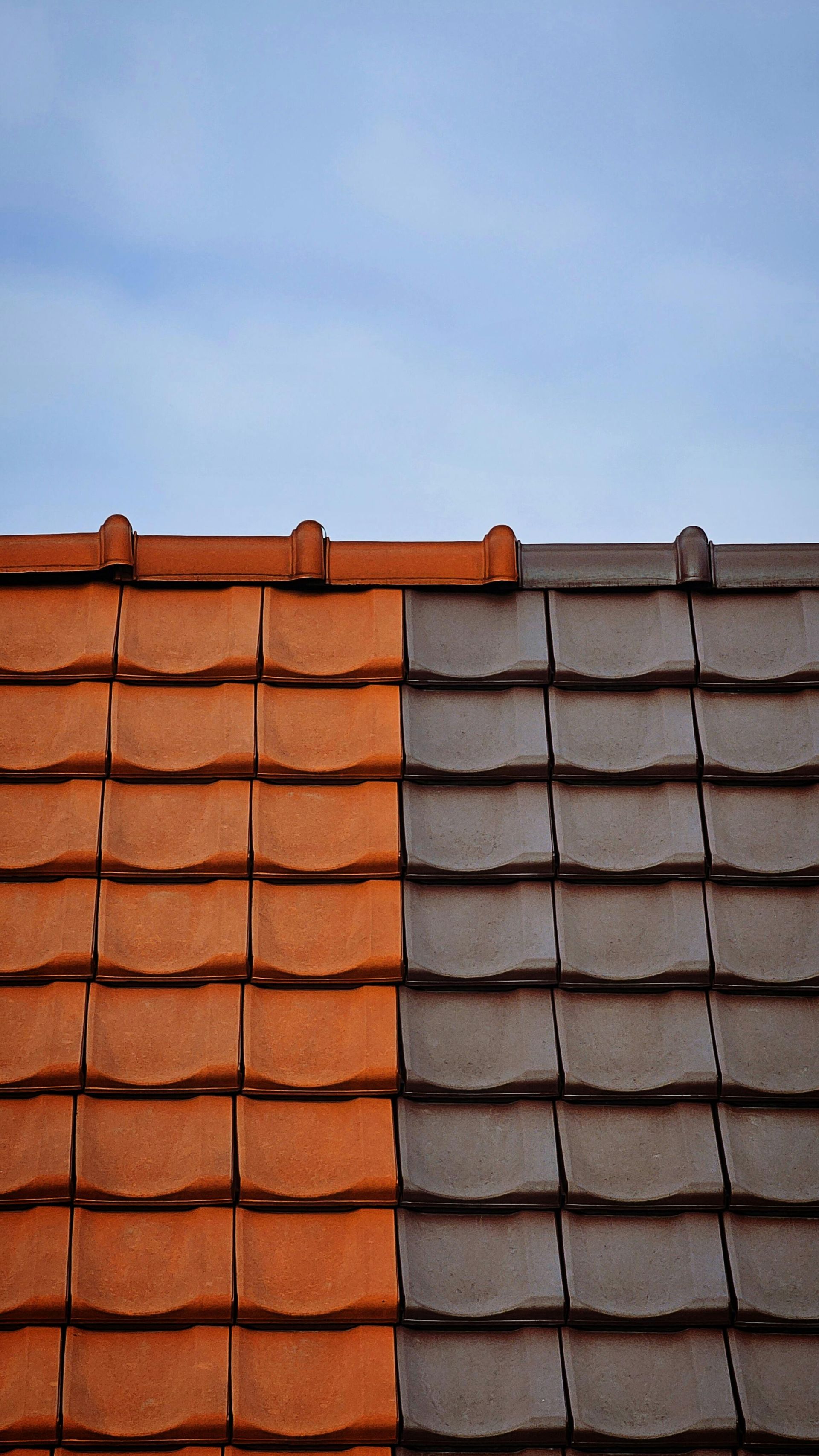 Two sections of a tiled roof, one orange and one brown, against a blue sky.