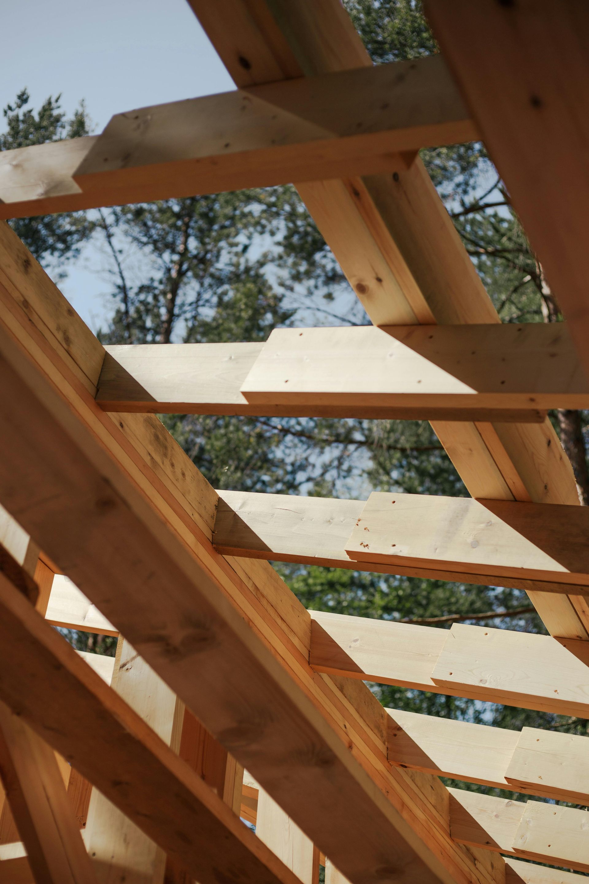 Two construction workers on a roof, installing wooden beams. One on ladder, one on ground. Blue sky background.